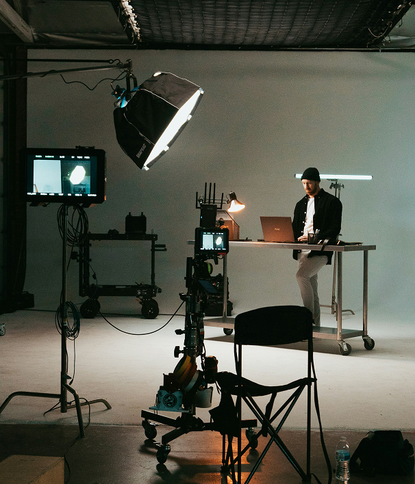 A film studio set with lighting, monitors and a camera, and a man working on a laptop at a steel table